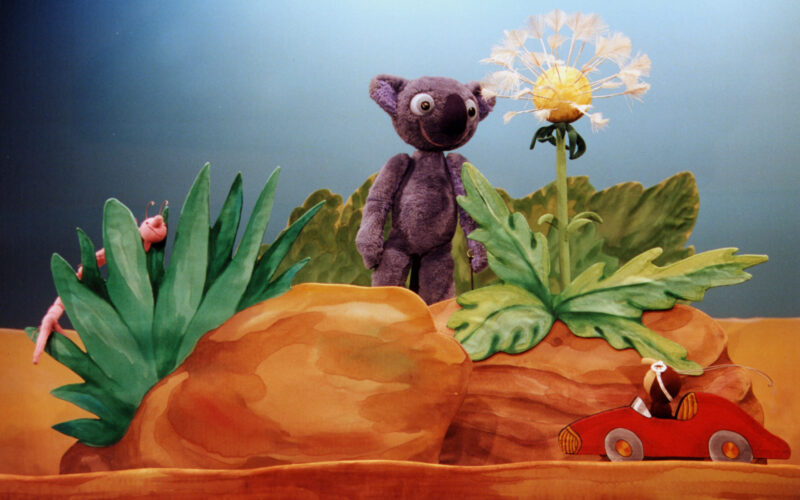 Ein lilafarbener Plüschbär steht neben einer Pusteblume zwischen Steinen, mit einem Spielzeugwurm auf einem Blatt und einer Maus in einem roten Spielzeugauto im Vordergrund.