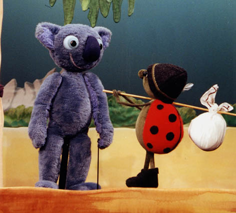 Eine blaue Koala-Puppe steht auf der Bühne neben einer Marienkäfer-Puppe, die einen Stock mit einem gebündelten Sack trägt.