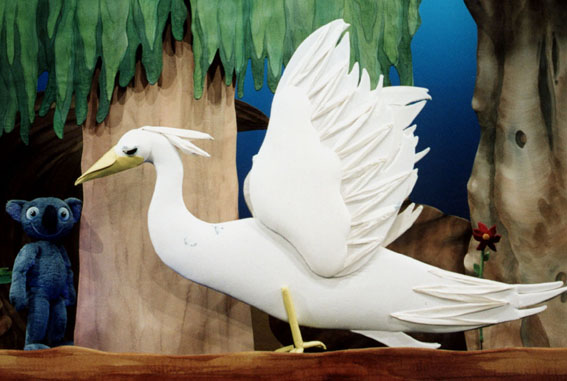 Ein weißer Puppenvogel mit erhobenen Flügeln steht auf einem Ast vor einer Kulisse mit Bäumen; im Hintergrund ist eine blaue teddybärähnliche Puppe zu sehen.