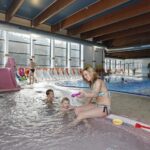 Ein Hallenbad mit Kindern und Erwachsenen; eine Frau sitzt mit zwei kleinen Kindern am Beckenrand, während andere in der Nähe schwimmen und spielen.