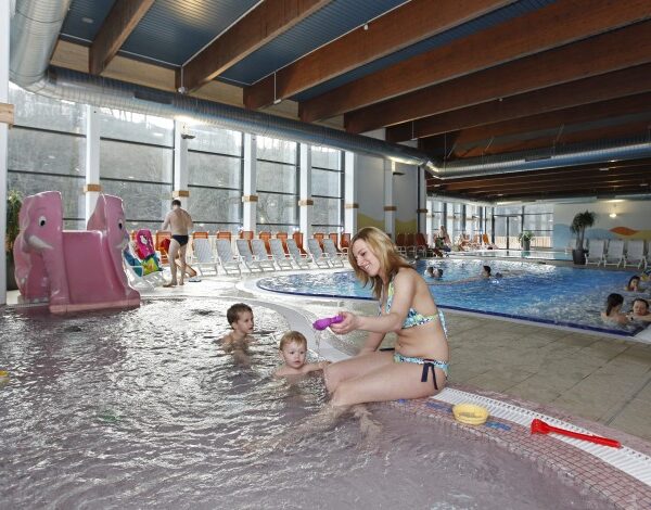 Ein Hallenbad mit Kindern und Erwachsenen; eine Frau sitzt mit zwei kleinen Kindern am Beckenrand, während andere in der Nähe schwimmen und spielen.