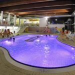 Hallenbad mit violetter Beleuchtung, mehrere Personen im Wasser, Liegestühle an den Seiten und große Fenster auf der rechten Seite.