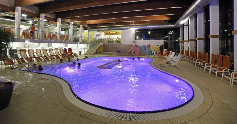 Hallenbad mit violetter Beleuchtung, mehrere Personen im Wasser, Liegestühle an den Seiten und große Fenster auf der rechten Seite.
