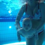 Ein Erwachsener im Badeanzug hält ein Baby unter Wasser in einem Schwimmbad. Beide sind untergetaucht, und im Hintergrund sind die Beckenfliesen zu sehen.