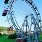 Ein großes Riesenrad mit roten Fahrgastkabinen steht auf einer Wiese unter einem strahlend blauen Himmel.