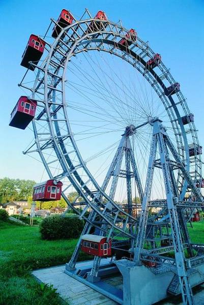 Ein großes Riesenrad mit roten Fahrgastkabinen steht auf einer Wiese unter einem strahlend blauen Himmel.