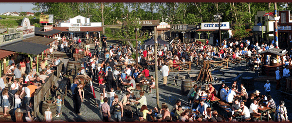 Eine große Menschenmenge versammelt sich bei sonnigem Wetter in einer Stadt im Freien, die dem alten Westen nachempfunden ist, mit Holzgebäuden und verschiedenen Schildern.