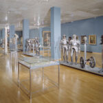 Museumsgalerie mit Exponaten von mittelalterlichen Rüstungen, Helmen und Waffen, die in Vitrinen und auf Ständern ausgestellt sind; blaue Wände und polierter Holzboden.