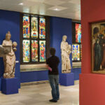 Eine Person steht in einer Museumsgalerie mit Statuen, Buntglasfenstern und einem Gemälde, das eine religiöse Szene an der Wand darstellt.