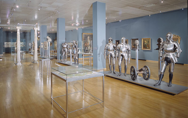 Museumsgalerie mit Glasvitrinen, mittelalterlichen Rüstungen, Helmen und historischen Artefakten, die auf Ständern vor blauen Wänden und einem polierten Holzfußboden aufgestellt sind.