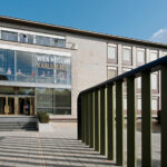 Außenansicht des Wien Museum Karlsplatz mit großen Glasfenstern, Eingangstreppe und grünem Geländer im Vordergrund an einem klaren Tag.