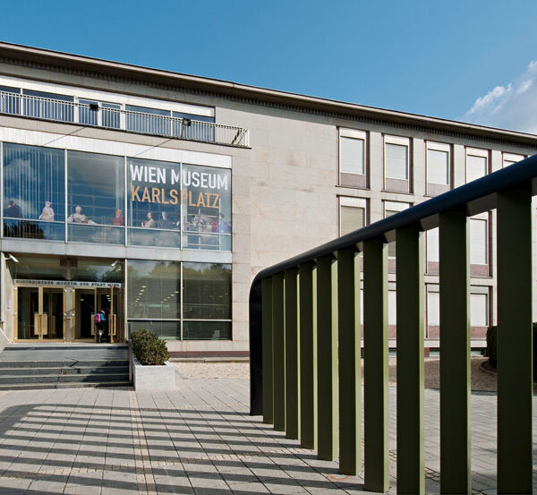 Außenansicht des Wien Museum Karlsplatz mit großen Glasfenstern, Eingangstreppe und grünem Geländer im Vordergrund an einem klaren Tag.