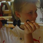 Zwei Jungen in weißen T-Shirts halten und untersuchen einen Metallstab mit schwarzen Griffen, umgeben von anderen Kindern in einem Haus.