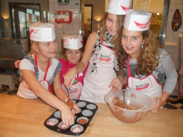 Vier Kinder mit Kochmützen und Schürzen aus Papier bereiten an einem Küchentisch gemeinsam Muffins zu und füllen den Teig aus einer Rührschüssel in ein Muffinblech.