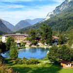 Ein kleiner See, umgeben von Bäumen, Gebäuden und Bergen unter blauem Himmel; im Vordergrund steht eine Holzhütte.