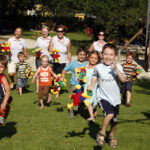 Eine Gruppe von Kindern rennt über eine Wiese und hält bunte Plüschtiere in der Hand, während im Hintergrund mehrere Erwachsene in der Nähe eines Spielplatzes lächeln.
