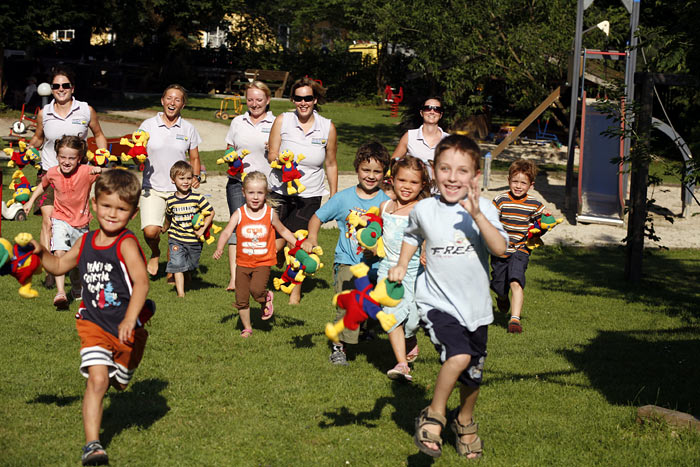 Eine Gruppe von Kindern rennt über eine Wiese und hält bunte Plüschtiere in der Hand, während im Hintergrund mehrere Erwachsene in der Nähe eines Spielplatzes lächeln.