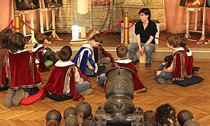 Eine Gruppe von Kindern in Kostümen sitzt auf einem Holzboden in einem Museum und hört zwei Erwachsenen zu, die vor Gemälden und einer historischen Kanone stehen.