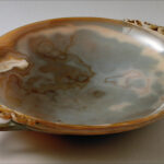 Eine flache, runde Schale aus Achat mit kunstvoll geschnitzten Henkeln auf jeder Seite, die natürliche Muster in Braun- und Grautönen aufweisen.