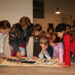 Eine Gruppe von Kindern und zwei Erwachsenen versammelt sich um einen Tisch, betrachtet und diskutiert ein buntes Kunstwerk, das vor ihnen ausgebreitet ist.