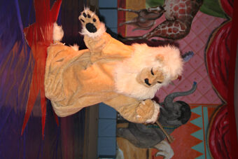 Person in einem Löwenkostüm, die auf der Bühne vor einem farbenfrohen Hintergrund mit Tiermotiven auftritt, darunter Gemälde einer Giraffe und eines Elefanten.