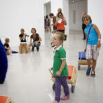 Kinder ziehen Holzkarren in einem hellen, modernen Galerieraum mit Erwachsenen und anderen Kindern im Hintergrund. Die Kunstwerke sind im ganzen Raum zu sehen.