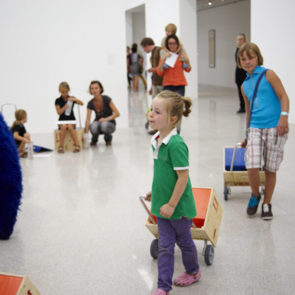 Kinder ziehen Holzkarren in einem hellen, modernen Galerieraum mit Erwachsenen und anderen Kindern im Hintergrund. Die Kunstwerke sind im ganzen Raum zu sehen.