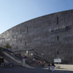 Ein großes, modernes, graues Steingebäude mit "mumok"-Schild; Menschen, die in der Nähe spazieren gehen und auf den Treppen am Eingang sitzen.