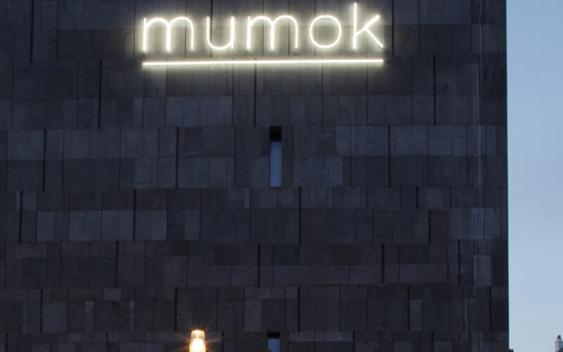 Ein dunkles Steingebäude mit beleuchtetem "mumok"-Schild, ein Museumseingang, draußen geparkte Fahrräder und eine leuchtende Straßenlaterne in der Abenddämmerung.