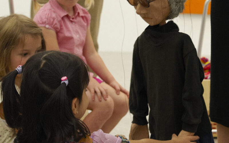 Eine Gruppe kleiner Kinder sitzt auf dem Boden und interagiert mit einer grauhaarigen Marionette, die von einem Erwachsenen in einer Galerie oder einem Klassenzimmer gesteuert wird.