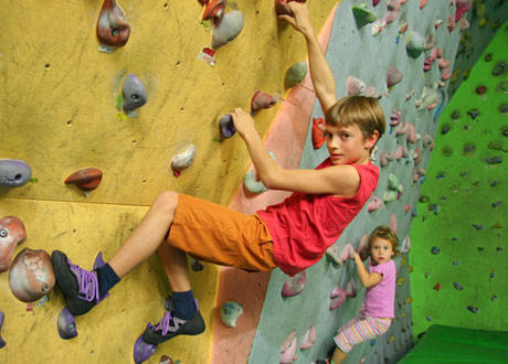 Zwei Kinder klettern an einer Boulderwand in einer Halle, wobei das ältere Kind im Vordergrund und das jüngere Kind dahinter steht.