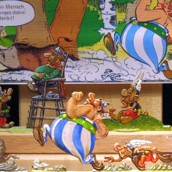 Diorama der Asterix-Comicfiguren mit Obelix, der einen Mann auf einem Stuhl trägt, vor dem Hintergrund einer Comictafel.