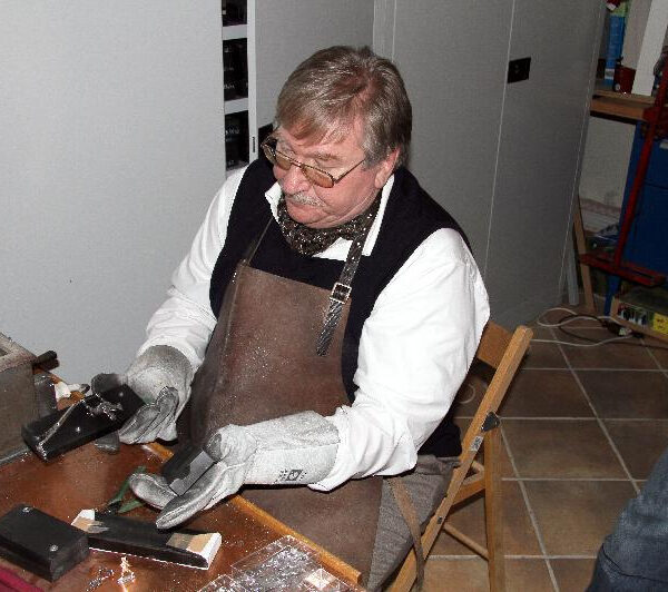 Ein Mann mit Handschuhen und Schürze sitzt an einer Werkbank, hantiert mit Werkzeugen und Materialien und scheint an einem kleinen Handwerks- oder Reparaturprojekt in einem Haus zu arbeiten.