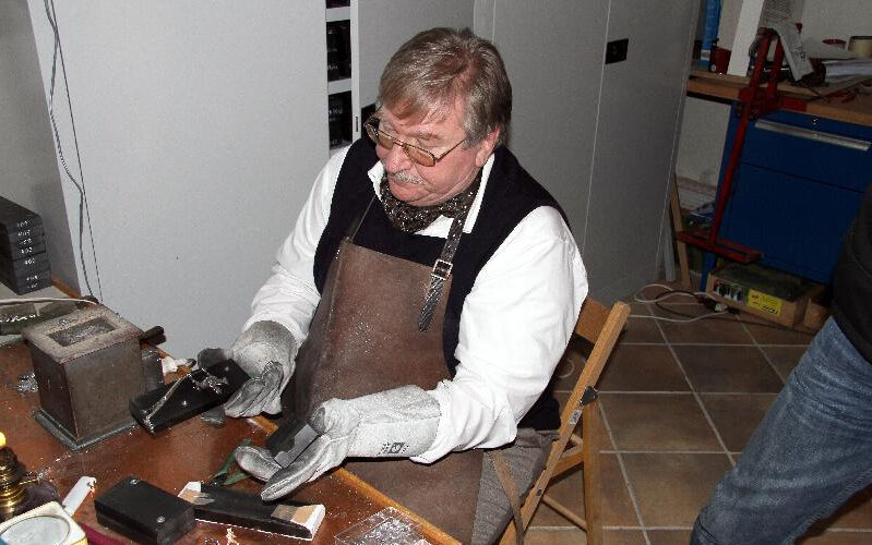 Ein Mann mit Handschuhen und Schürze sitzt an einer Werkbank, hantiert mit Werkzeugen und Materialien und scheint an einem kleinen Handwerks- oder Reparaturprojekt in einem Haus zu arbeiten.