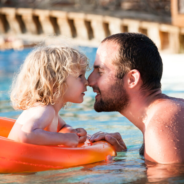 Ein kleines Kind in einem orangefarbenen Schwimmkörper und ein erwachsener Mann berühren sich lächelnd die Nasen, während sie in einem Schwimmbad sind.