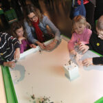 Ein Erwachsener und vier Kinder interagieren mit kleinen Objekten auf einem weißen Tisch in einem hell erleuchteten, modernen Ausstellungsraum.