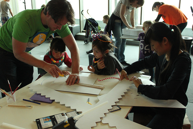 In einem hell erleuchteten Raum sitzen Erwachsene und Kinder an einem Tisch und arbeiten mit Papier, Lineal und Schere an Bastelprojekten.