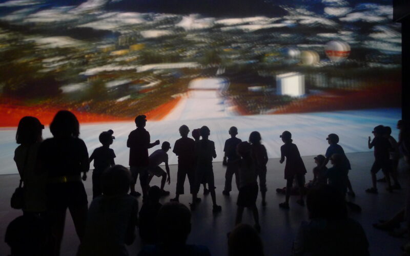 Eine schemenhafte Gruppe von Kindern und Erwachsenen steht und sitzt vor einer großen, verschwommenen Projektion einer Landschaftsszene.