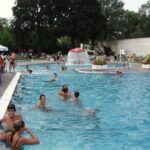 Der Außenpool mit Wasserfontäne, Rutsche und Liegestühlen, umgeben von Bäumen und Grün, lädt zum Schwimmen und Entspannen ein.