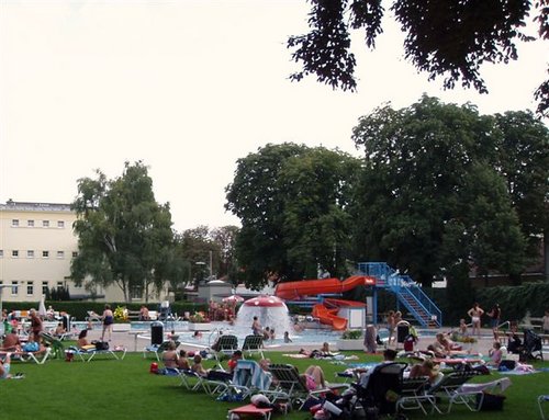Man kann sich auf Liegestühlen entspannen und in einem öffentlichen Freibad mit roter Rutsche, Wasserspielen und umliegenden Bäumen schwimmen.
