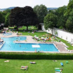 Ein großer Freibadkomplex mit Rutsche, umgeben von Gras, Bäumen und Menschen, die sich sonnen oder auf Liegestühlen entspannen.