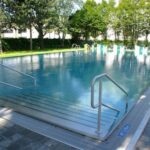 Außenschwimmbad mit Stufeneinstieg und Handläufen, umgeben von Bäumen und einer gepflasterten Fläche; im Hintergrund sind Schwimmbadschilder und Stühle zu sehen.