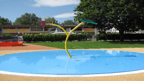 Ein kleines Planschbecken mit seichtem, blauem Wasser, zwei gelbe Sprühvorrichtungen in Form von Blättern und ein Schulgebäude im Hintergrund an einem sonnigen Tag.