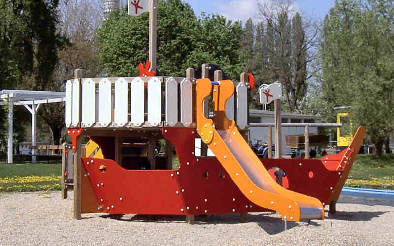Eine Spielplatzstruktur in Form eines rot-weißen Schiffes mit einer orangefarbenen Rutsche, die auf einer Kiesfläche im Freien steht.