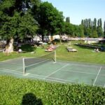 Badmintonplatz im Freien auf einer Wiese mit Sonnenbadenden und Entspannern auf Liegestühlen im Hintergrund, umgeben von Bäumen.