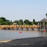Ein hölzerner Steg mit Geländer überquert ein ruhiges Gewässer und führt zu einem Pavillon auf der rechten Seite. Im Hintergrund sind Bäume und Gebäude zu sehen.