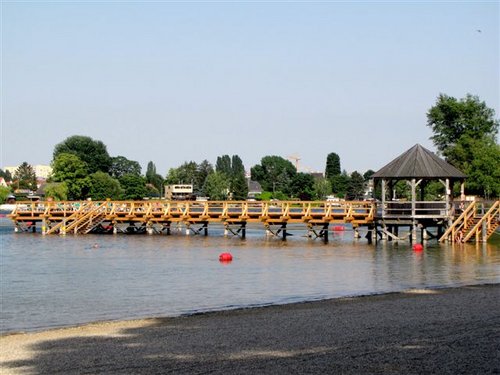 Ein hölzerner Steg mit Geländer überquert ein ruhiges Gewässer und führt zu einem Pavillon auf der rechten Seite. Im Hintergrund sind Bäume und Gebäude zu sehen.