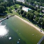 Luftaufnahme eines Strandes am See mit schwimmenden und sich sonnenden Menschen; in der Nähe befinden sich Grasflächen, ein Pier, ein Pool und geparkte Autos.