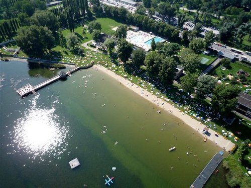 Luftaufnahme eines Strandes am See mit schwimmenden und sich sonnenden Menschen; in der Nähe befinden sich Grasflächen, ein Pier, ein Pool und geparkte Autos.