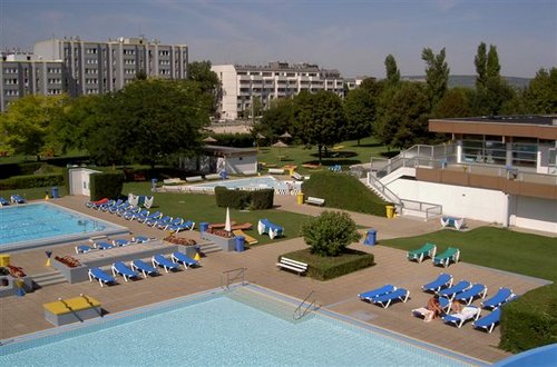 Freibadkomplex mit mehreren Becken, zahlreichen blauen Liegestühlen, ein paar Sonnenbadenden und Wohngebäuden im Hintergrund.